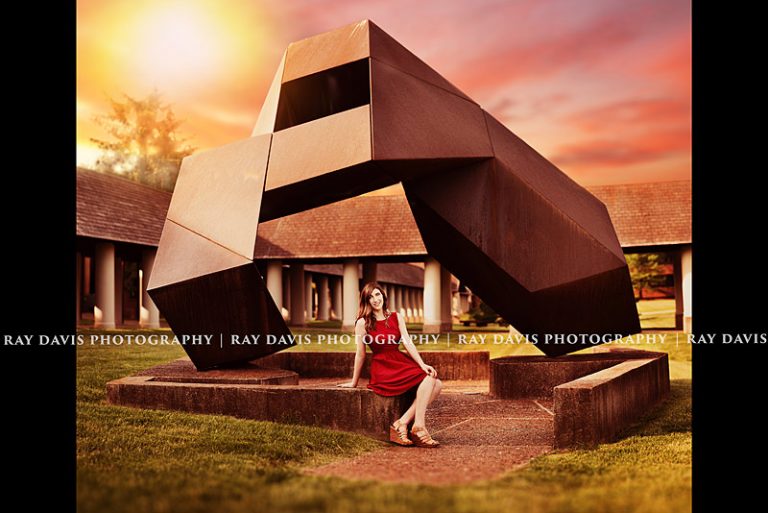 Grad sits under Bellarmine University Campus sculpture in red dress by Ray Davis Photography